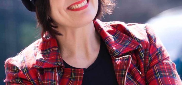 “Children Are The World’s Play Experts” Virginia wears a black baker-boy hat over cropped brown hair, with a red tartan jacket and black tshirt. Her arms are crossed and she is smiling in the sunshine.